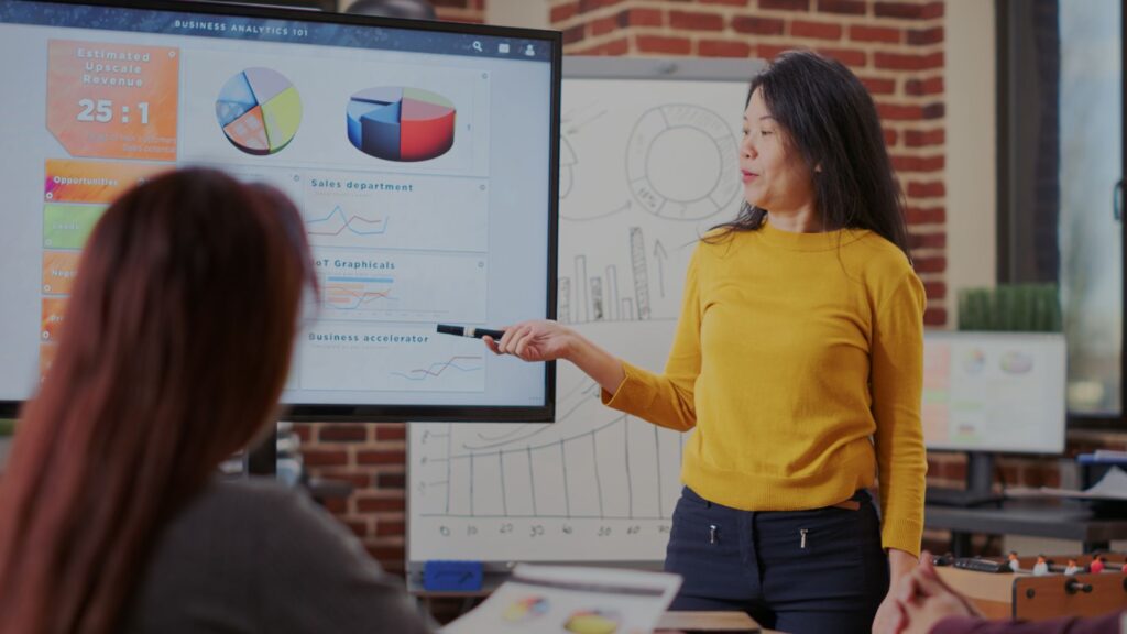 A woman in a yellow sweater offers practical guidance as she points to charts on a large screen during a business presentation in a modern office.