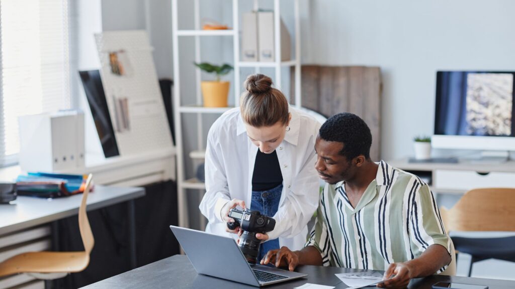 A woman examines a camera while a man looks at a laptop on a desk in a bright, modern office setting, exploring recommended animation services for their next creative project.