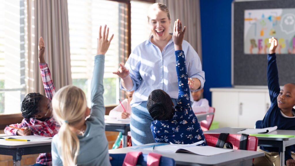 A teacher stands at the front of a classroom smiling as several students seated at desks raise their hands to answer a question, demonstrating effective teacher training in action.