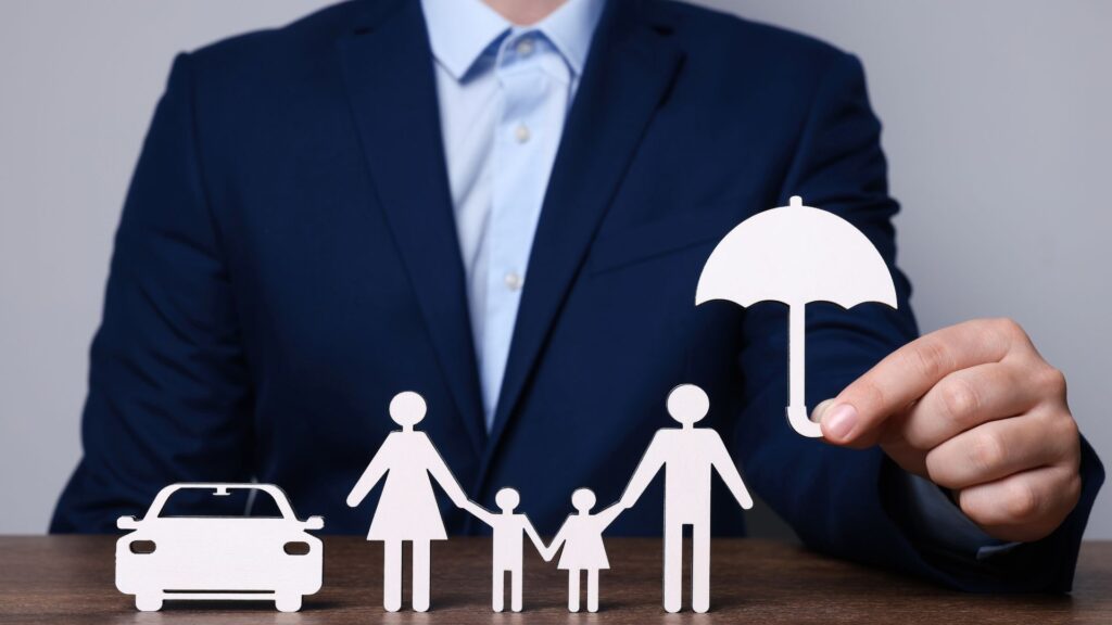 A person in a suit holds a paper umbrella over cutouts of a car and family, visually conveying the insurance animation benefits of protection and security.