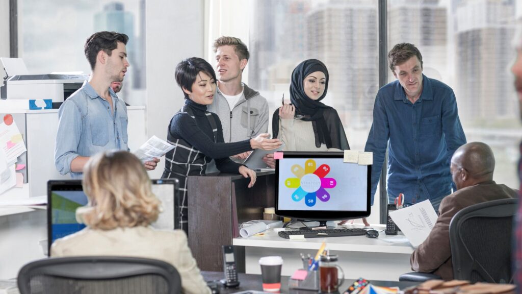 A group of six coworkers have a discussion around a desk in a modern office, while a computer screen displays a colorful Reduce Training Costs Animation.