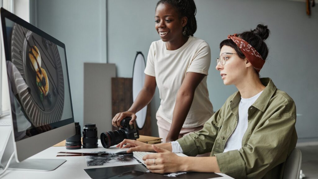 Two women working at a computer desk with a camera and lenses, reviewing a food photograph on a large monitor as they discuss AI solutions for interactive animation and editing techniques.