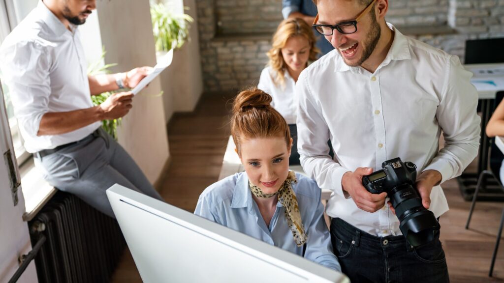 Two colleagues look at a computer screen in an office, discussing urgent animation services; one holds a camera while others work in the background.
