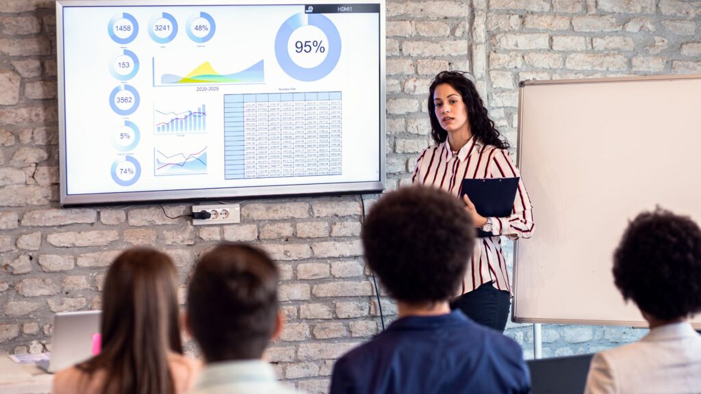 A woman uses Animation for PowerPoint to present data charts on a large screen to a group of seated people in a classroom with a brick wall and blank whiteboard.