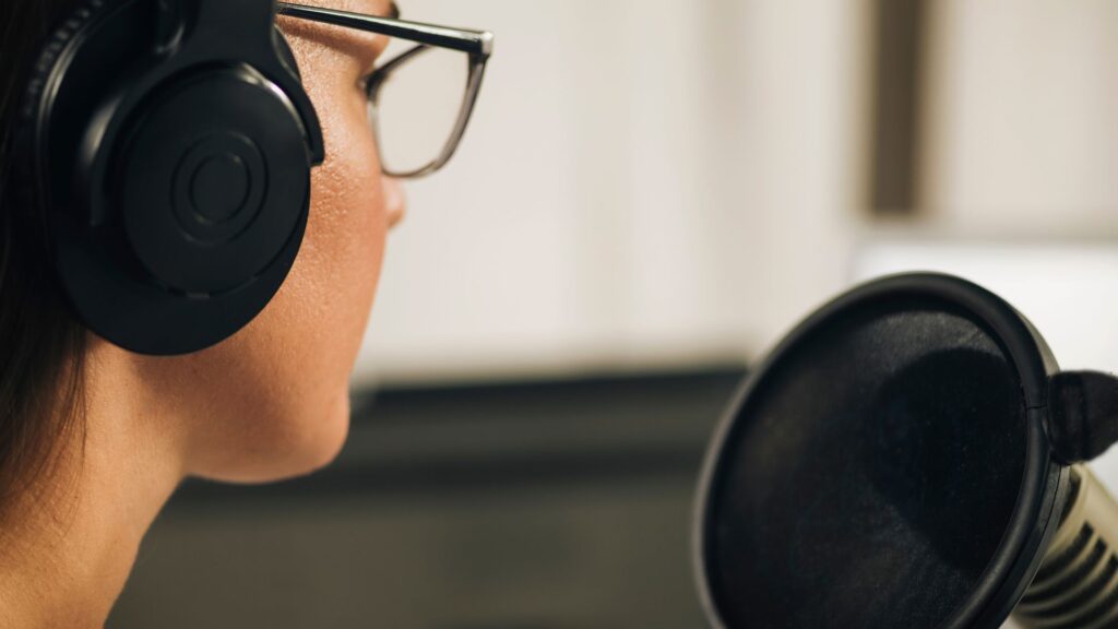 Close-up of a person wearing glasses and headphones, speaking into a microphone in a recording or broadcasting setup, ready to deliver urgent animation services with professional clarity.