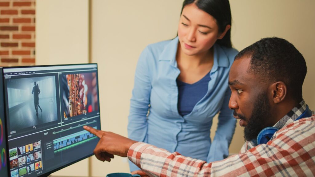Two people review and discuss video clips displayed on a computer monitor in an editing software interface, exploring ways to reduce training costs with engaging animation.