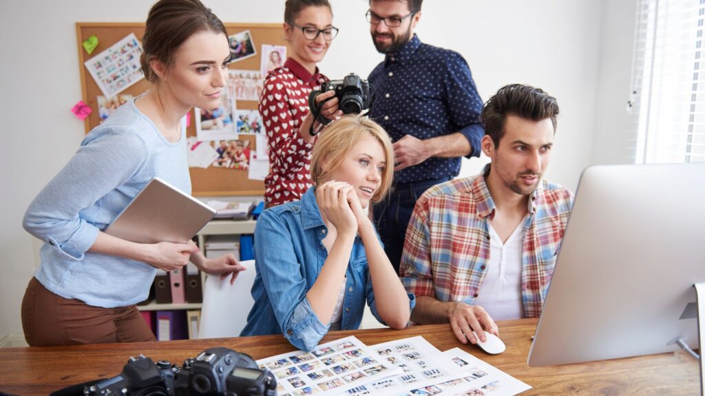 Five people collaborating at a desk, focused on a computer screen with printed photos and cameras nearby in a bright office—working intently on the Rush Animation Project.