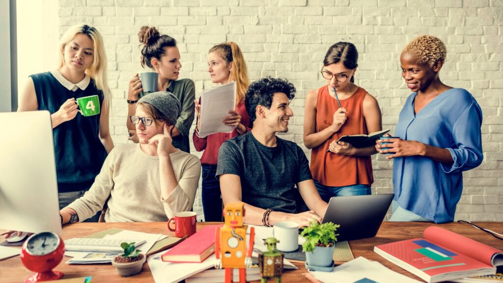 Seven people collaborate in a modern office, gathered around a desk with laptops, notebooks, and coffee mugs, brainstorming ideas for an explain services animation against a white brick wall background.