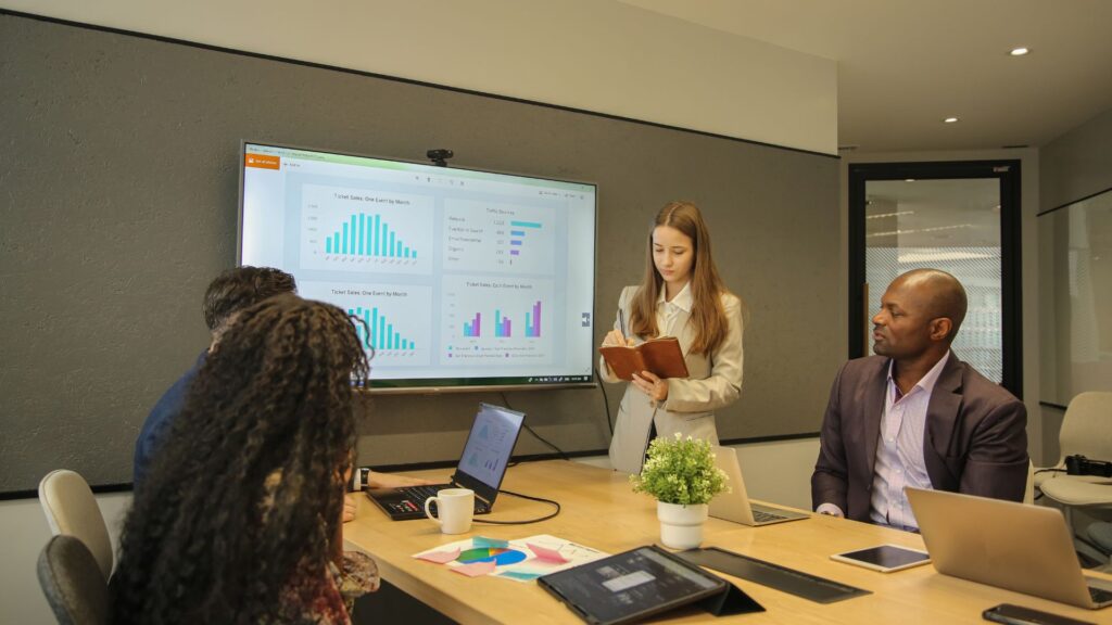Four people in a meeting room look at charts displayed on a large screen with Animation for PowerPoint, while one woman stands and takes notes; laptops and documents are on the table.