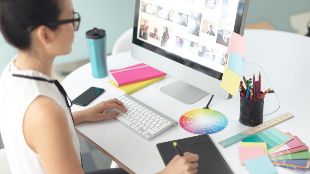 A person sits at a desk working on a computer displaying image thumbnails, surrounded by notebooks, colorful sticky notes, stationery, a color wheel, and materials for Enterprise Animation Solutions.