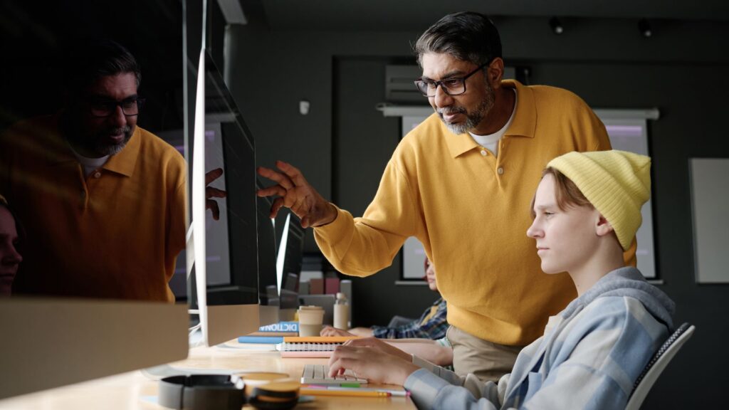 An adult wearing glasses and a yellow sweater points at a computer screen displaying a Reduce Training Costs animation, while a young person in a yellow beanie types on a keyboard in an office setting.