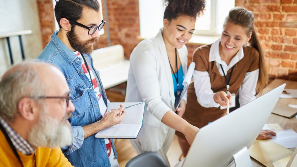 Four people gather around a computer in an office, discussing and taking notes about SME Animation Services, with one person pointing at the monitor.