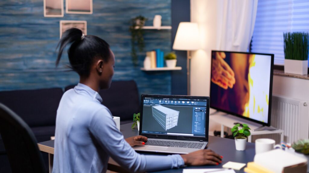A person sits at a desk working on a 3D modeling program on a laptop, exploring Enterprise Animation Solutions, with a TV screen and various office items visible in a modern home office setting.