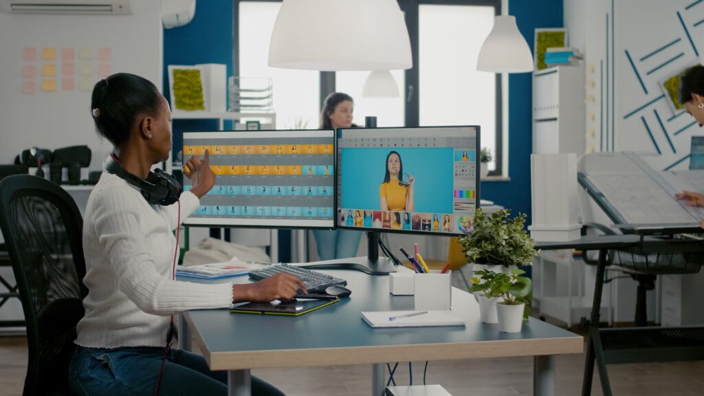 Woman sits at desk with dual monitors, editing photos of a person in yellow for a Priority Animation Project on one screen and sorting files on the other, in a modern office setting with colleagues in the background.