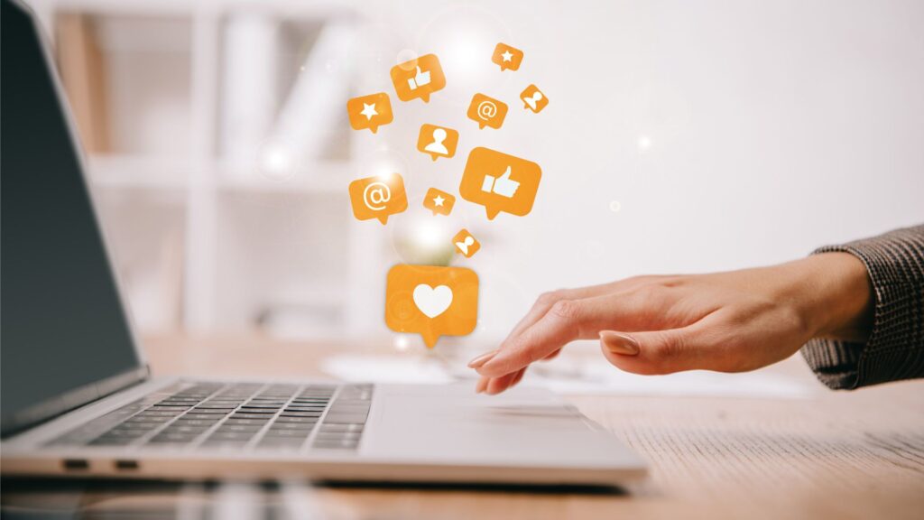 A hand uses a laptop touchpad with illustrated social media icons—hearts, likes, and comments—appearing above the keyboard, capturing the essence of Social Media Animation Services in action.