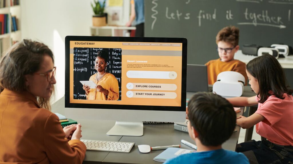 A teacher and students sit at a table with laptops and tablets; a large monitor displays an engaging learning experience on an online education platform with a teacher on the screen.