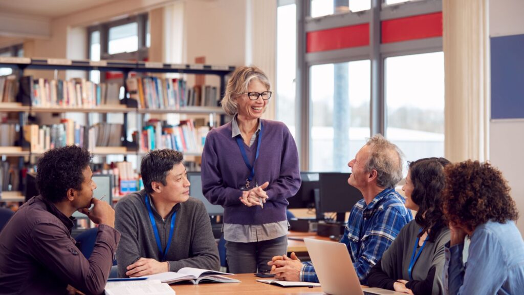 A group of adults sits around a table in a library, listening to a woman from Educational Voice standing and speaking, with books and laptops on the table.