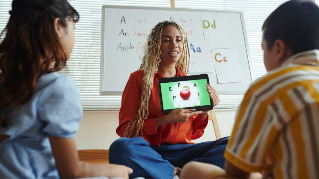 A woman shows two children a tablet with an engaging learning animation of an apple and letters, sitting in a classroom with alphabet posters on the whiteboard behind them.