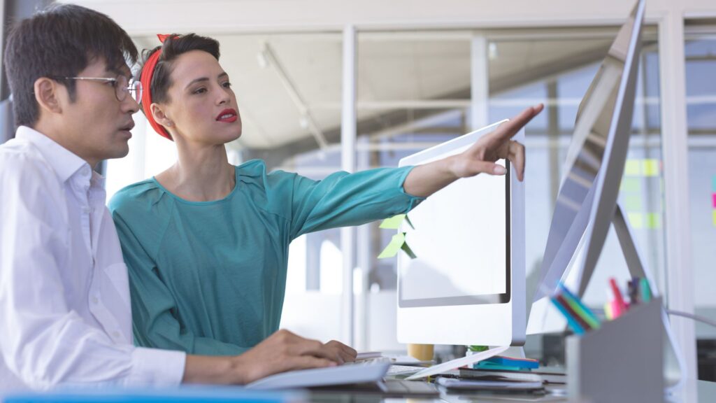 Two people sit at a desk looking at a large computer monitor; one person points at the screen while the other types on a keyboard, collaborating on an animation project under £1000.