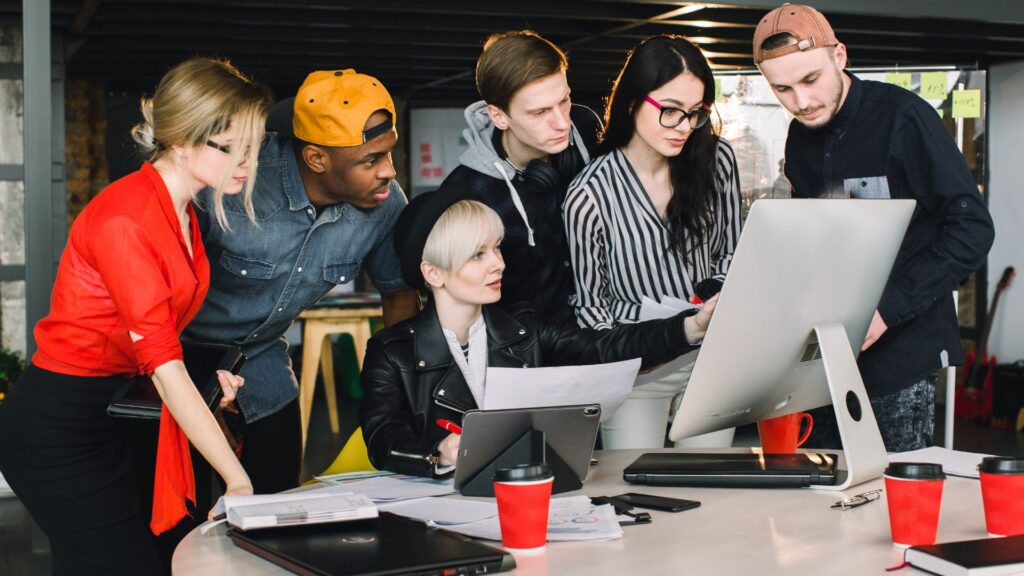 Six young adults gather around a desk, looking at a large computer monitor as they review documents and discuss an explain services animation in a modern office setting.