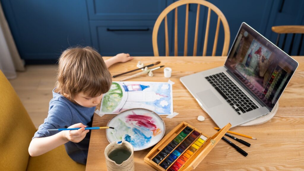 A child paints with watercolors at a table, surrounded by art supplies and a drawing, while their laptop streams an engaging online course animation as part of their art lesson.