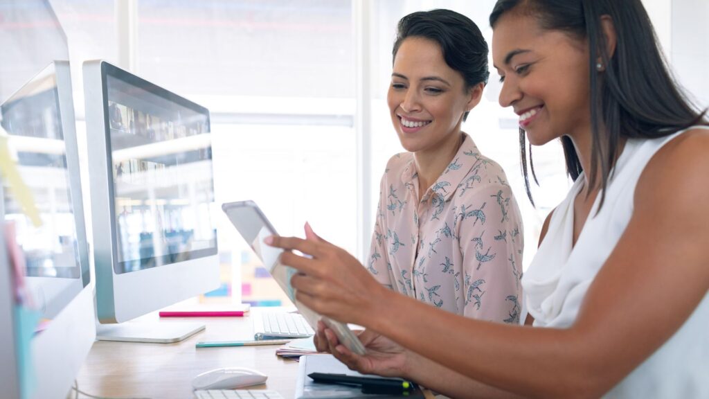 Two women sit at a desk with computers, smiling and looking at a tablet together in a bright office setting, exploring SME Animation Services.