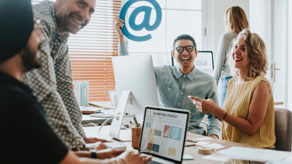 Four people sit around an office table, smiling and talking. One person holds up a large "@" symbol, discussing ideas for animation for email marketing. Laptops and papers are spread out on the table.