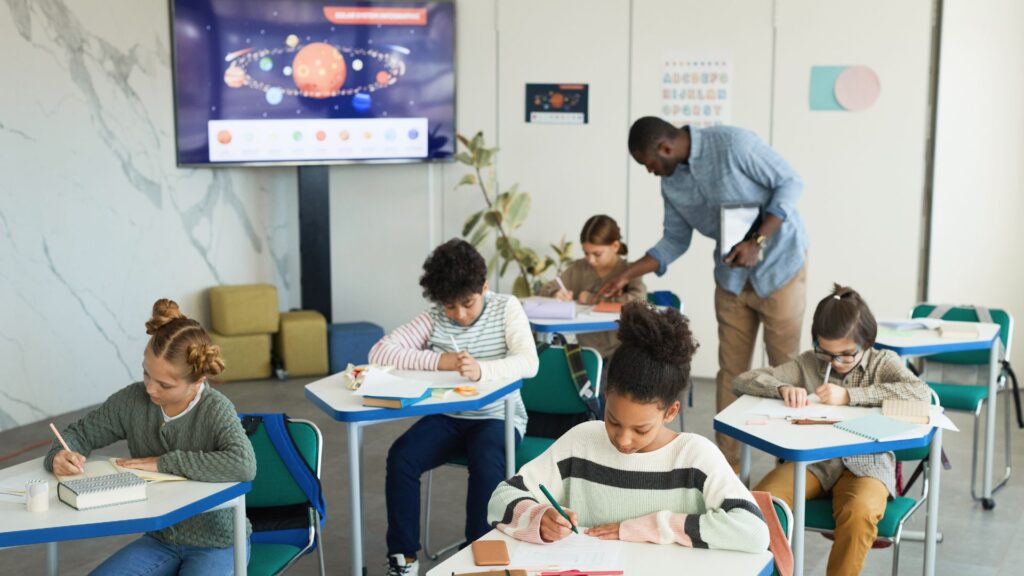 A teacher assists a group of elementary students working at desks in a classroom, using an engaging learning approach as a solar system diagram is displayed on a screen at the front.