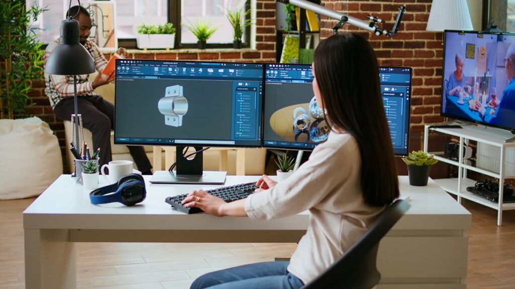 A woman works at a desk with dual monitors displaying 3D modeling software, using animation to simplify technical concepts in a modern home office, while a man sits in the background and a TV shows a cooking show.