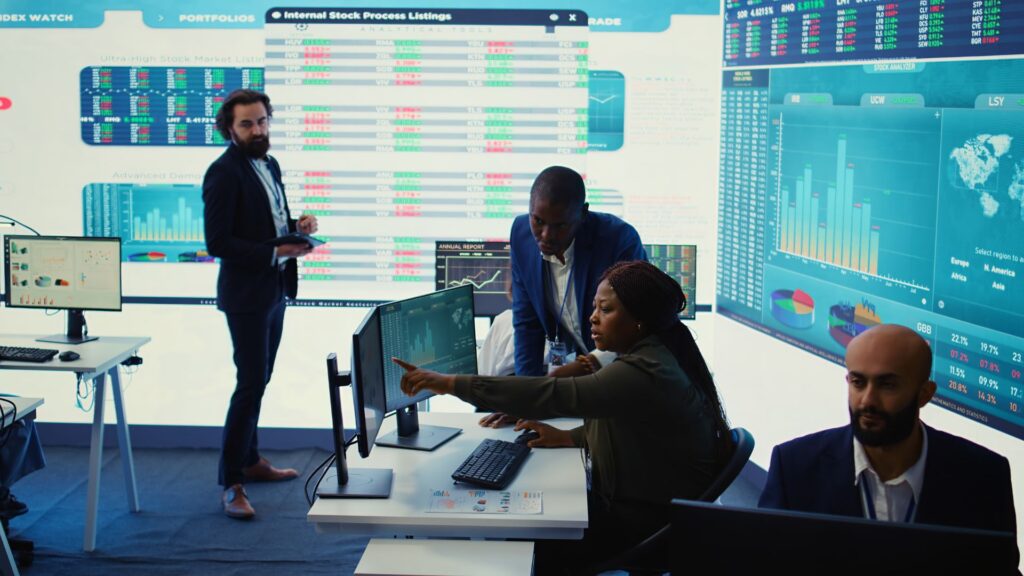 Four people analyze financial data on large screens in a modern office. One woman points at a monitor while others review charts, stock market information, and Banking Videos.