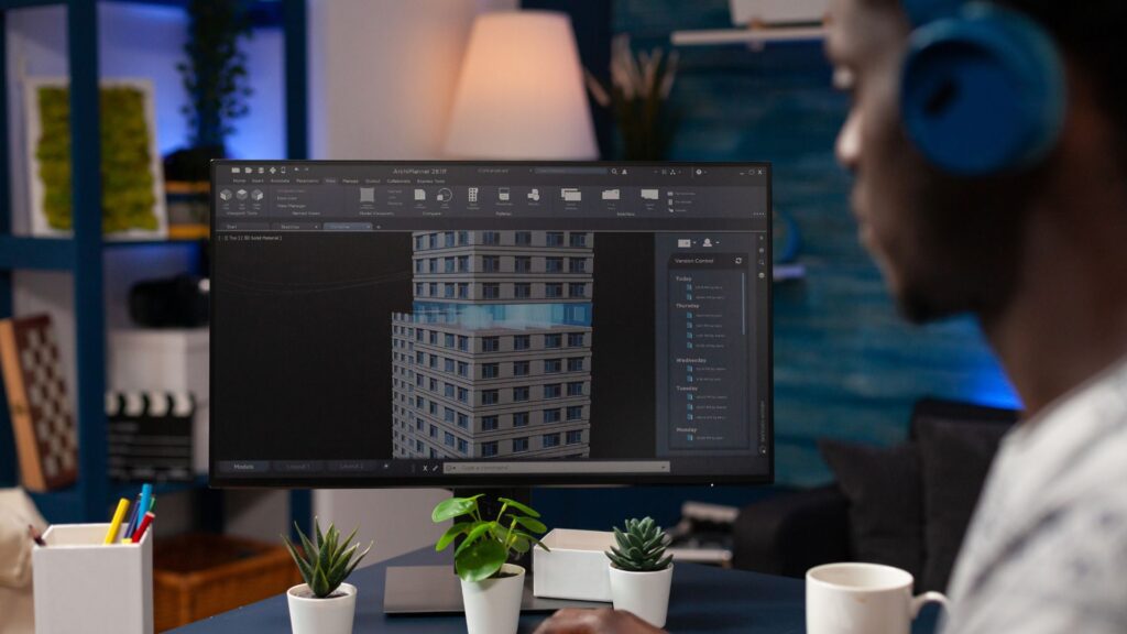 A person wearing headphones works on a 3D building model displayed on a computer monitor, using animation to simplify technical concepts, with small potted plants and office supplies on the desk.