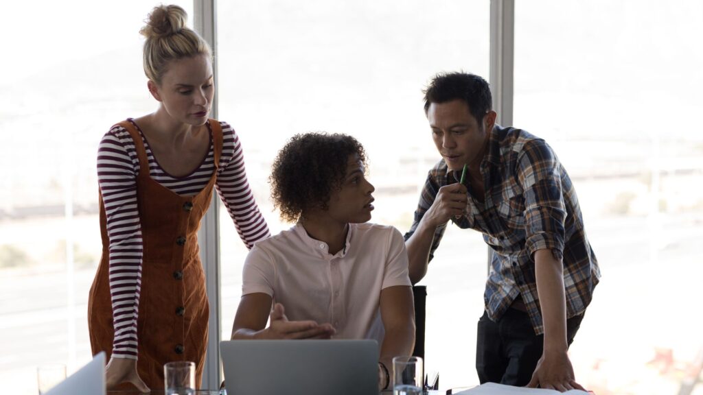 Three people have a discussion around a laptop in an office setting, exploring Emergency Animation Services, with papers and glasses of water on the table and large windows in the background.