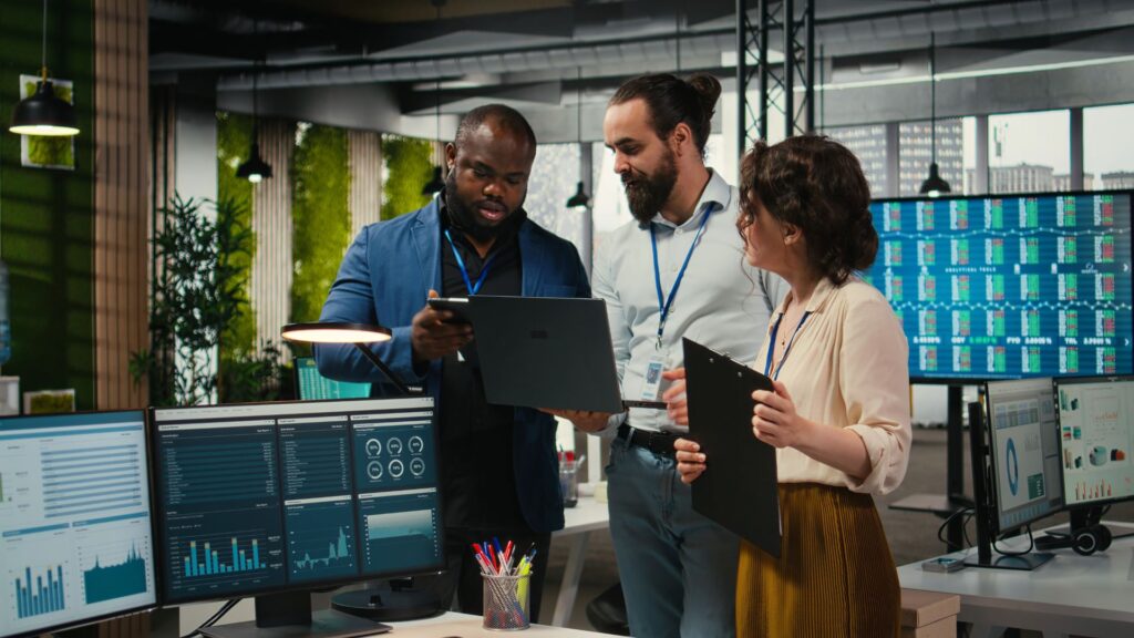 Three office workers discuss data displayed on a laptop in a modern workspace, with multiple computer screens showing charts, graphs, and Banking Videos.