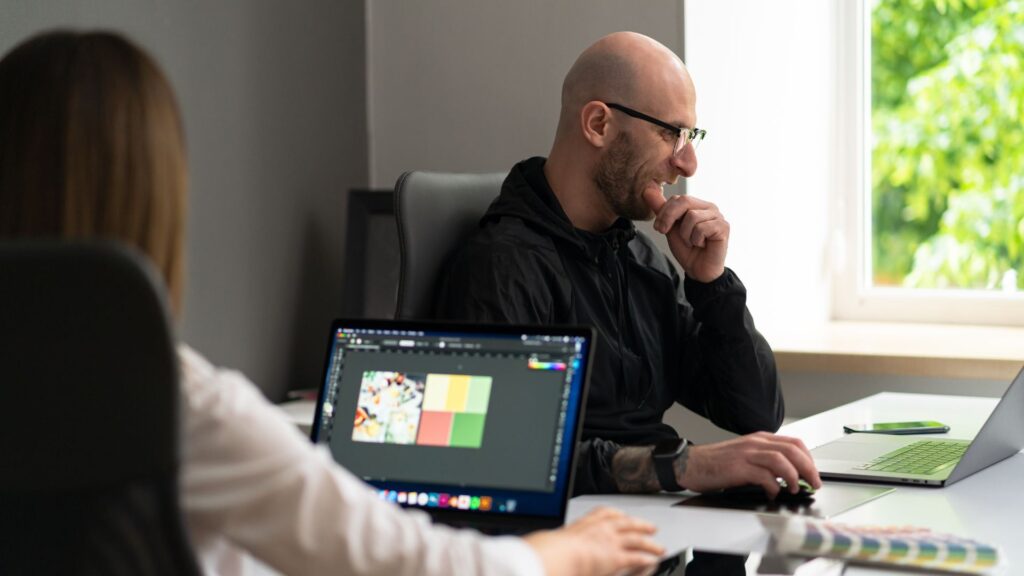 A man sits at a desk working on a laptop, appearing thoughtful. Nearby, a woman reviews fast animation production techniques on her colorful laptop screen. Lush greenery is visible through the window in the background.