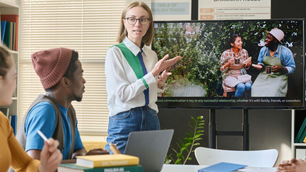 A woman presents to a group, gesturing at a screen displaying two people eating and talking, with an engaging caption about the importance of good communication in classroom learning.