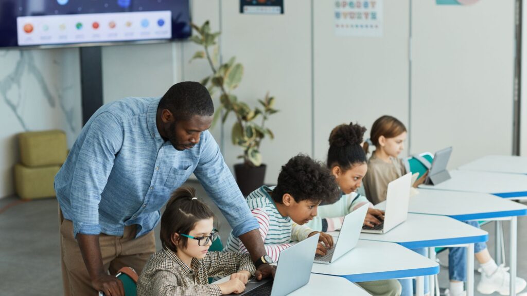 A teacher helps a student with a laptop while other students work at their desks, creating an environment of engaging classroom learning enhanced by technology.