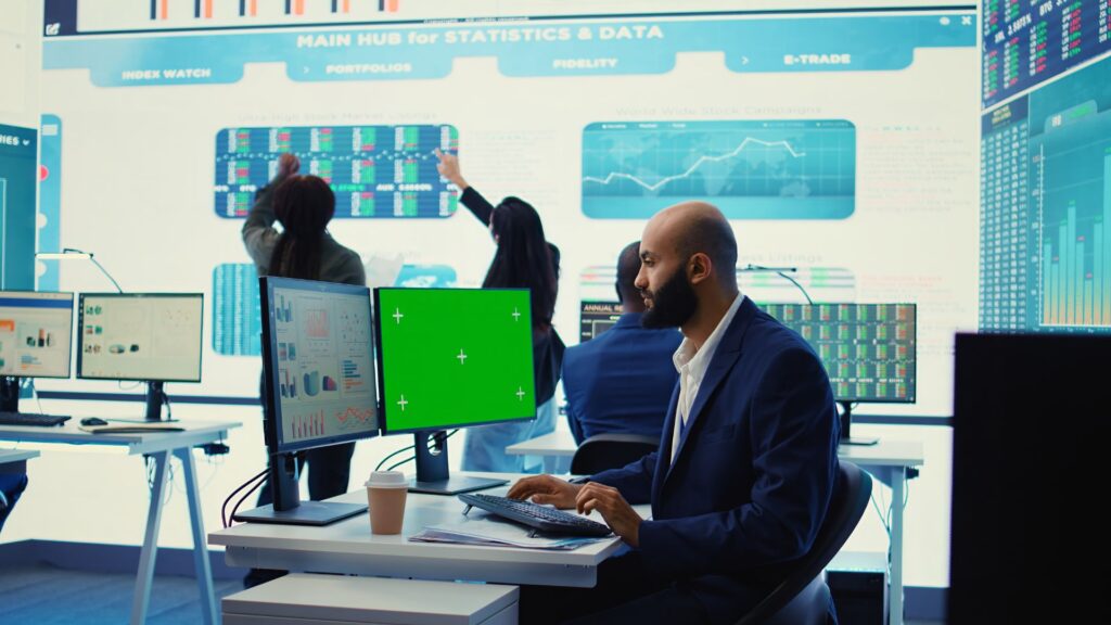 Man in a suit works at a desk with dual monitors in a busy office, while colleagues analyze Banking Videos and data on large digital screens in the background.