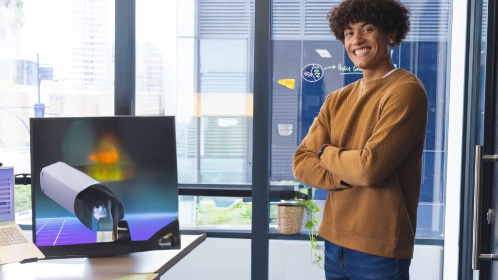 A person stands smiling with arms crossed beside a monitor displaying a 3D rendering of a satellite, showcasing Small Business Animation Services in a modern office setting.