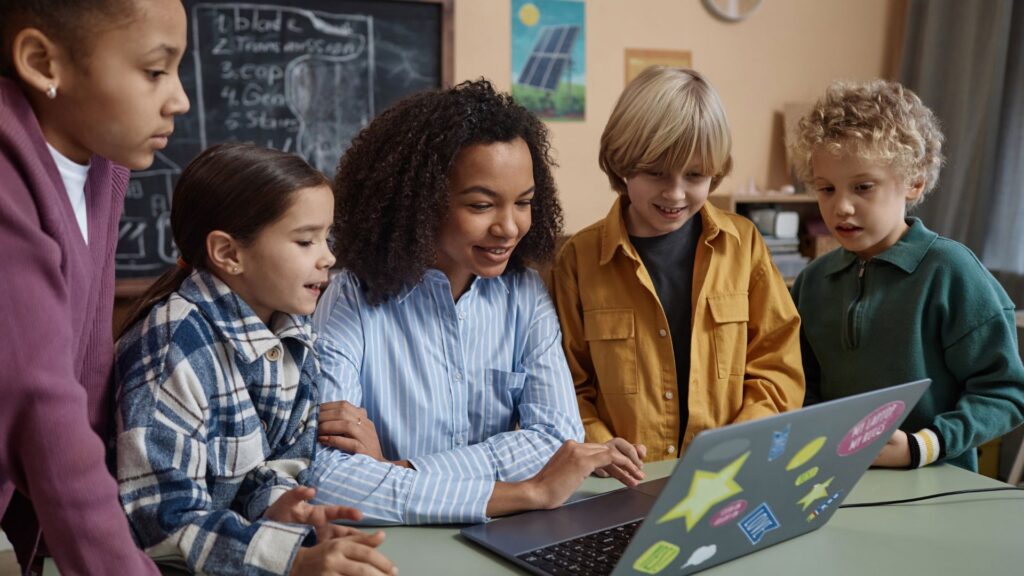 A group of five children gathers around a laptop with colorful stickers, engaging in classroom learning as they watch curriculum animation videos together in a vibrant classroom setting.