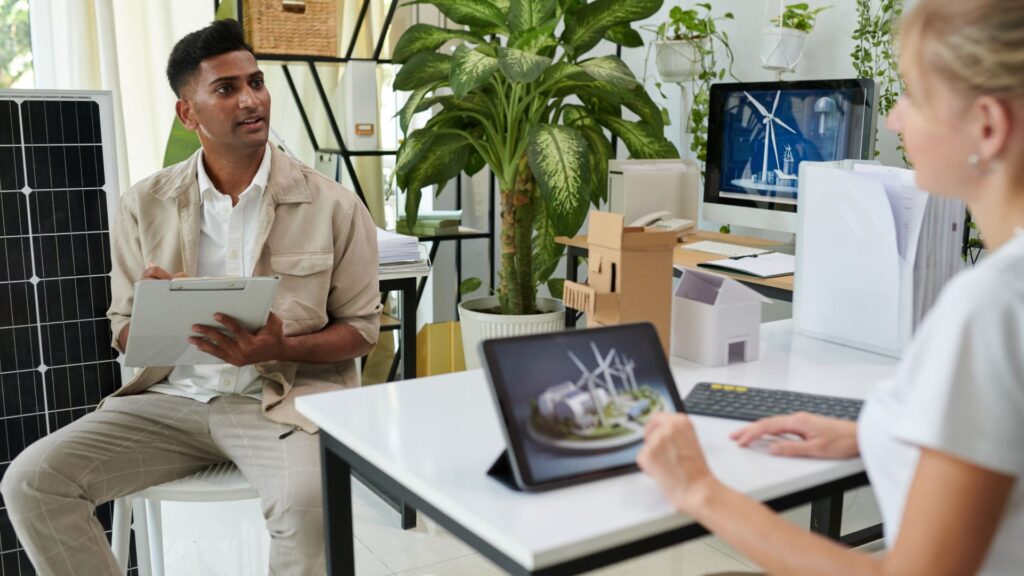 A man holding a clipboard talks to a woman at a desk with a tablet displaying wind turbines; plants, solar panel, and Small Business Animation Services branding in the background suggest an eco-friendly office.