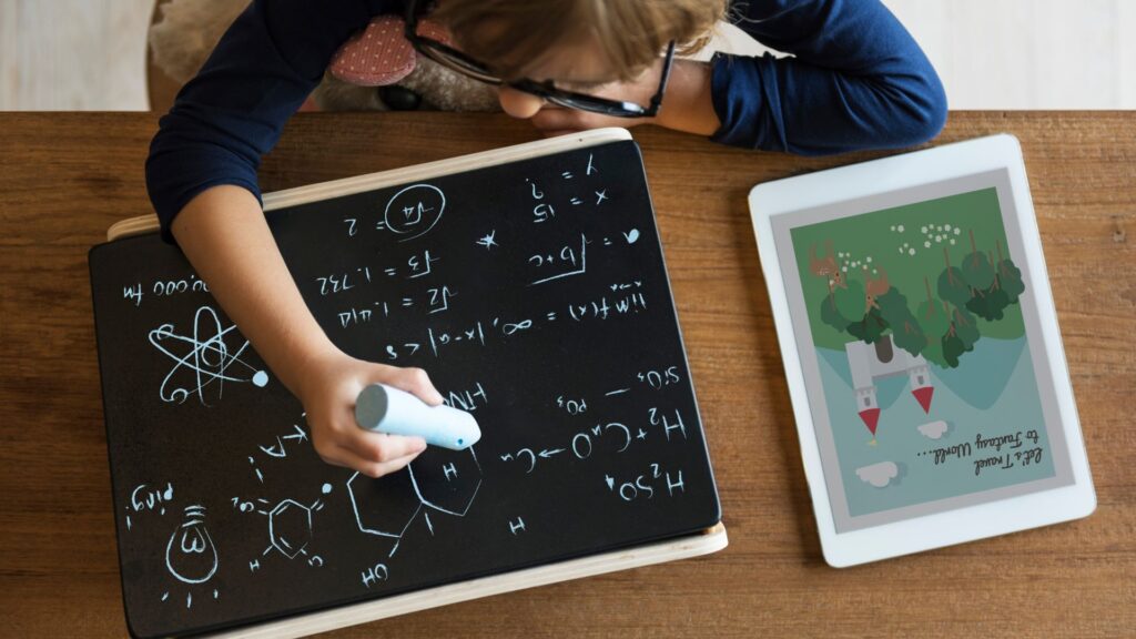 A child writes chemistry equations on a chalkboard with a tablet displaying engaging Curriculum Animation Videos of trees and a windmill on the table beside them, enriching their classroom learning experience.