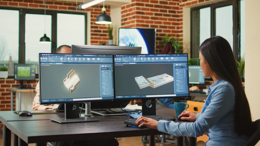 A woman works at a desk using two monitors displaying 3D CAD models in an office with brick walls and large windows—an inspiring scene for animation agencies near me seeking creative professionals.