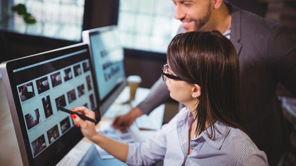 A woman points at images on a computer monitor with a pen while a man stands beside her, both reviewing Expedited Animation Services in an office setting.