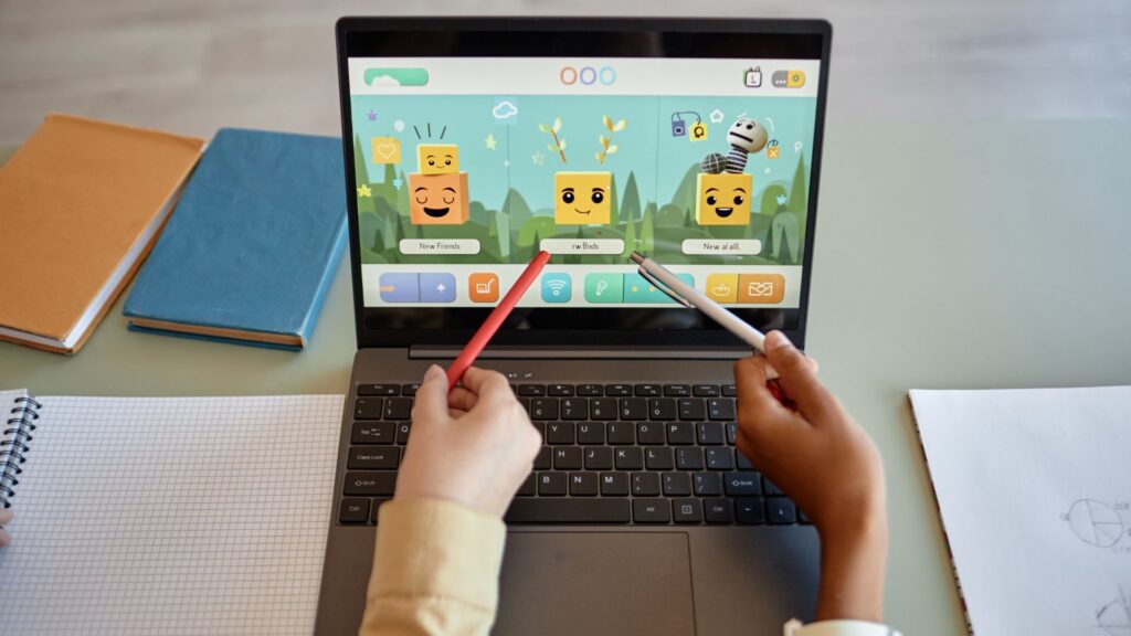 Two people point colored pens at a laptop screen displaying an engaging educational game with animated characters and text options. Books and a notepad sit nearby, creating a lively atmosphere for classroom learning.