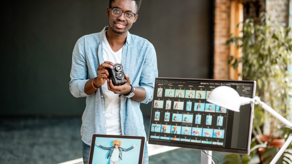 Man holding a camera stands beside a desk with a monitor and laptop displaying photo editing software and image thumbnails—ideal for creators seeking Expedited Animation Services.