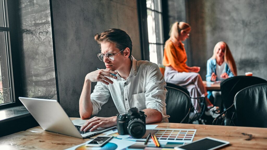 A man works on a laptop at a table with a camera, color swatches, and papers—possibly discussing Expedited Animation Services—while two women talk near a window in the modern cafe.