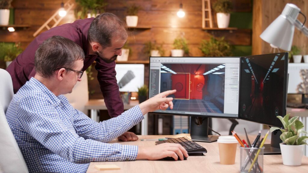 Two men in an office review 3D renderings of a red room on dual monitors; one is seated and pointing at the screen while the other stands and leans over his shoulder, discussing Small Business Animation Services.