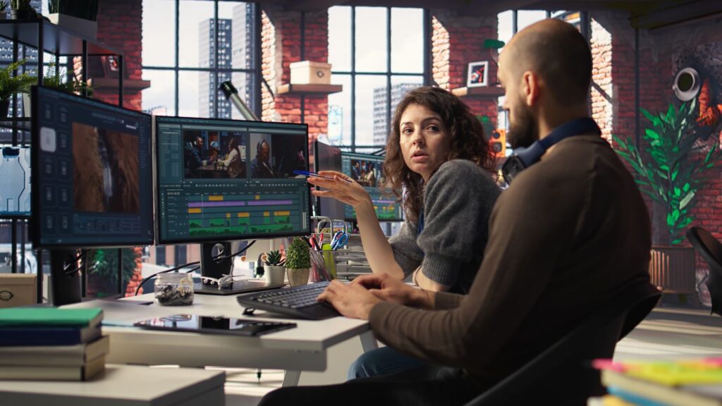 Two people sit at a desk with dual monitors, discussing how to improve engagement with an animated video project in a modern office with large windows and city views.