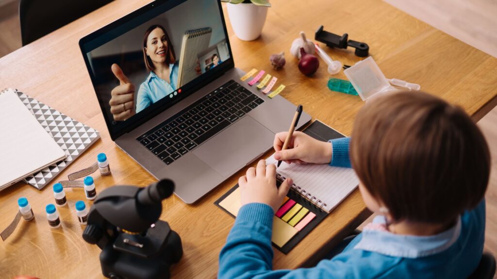 A child writes in a notebook during an engaging online lesson on a laptop, surrounded by science equipment and colored sticky notes on the wooden desk.