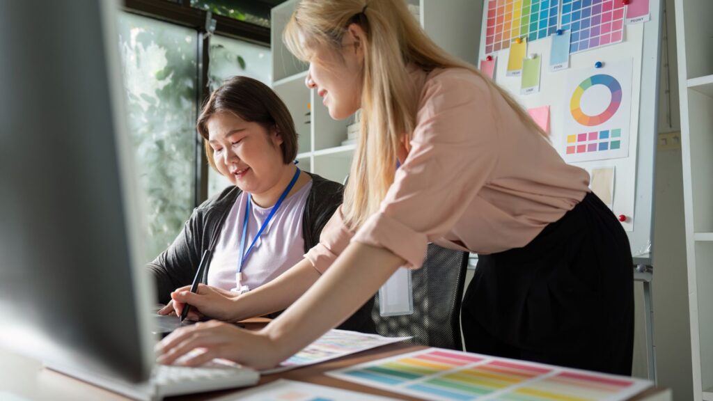 Two women collaborate at a desk with a computer, color swatches, and design charts in a well-lit office, discussing ideas for Expedited Animation Services.